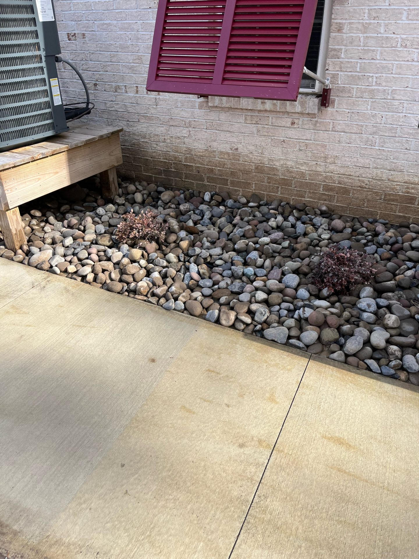Air conditioner near pebbled garden and brick wall.
