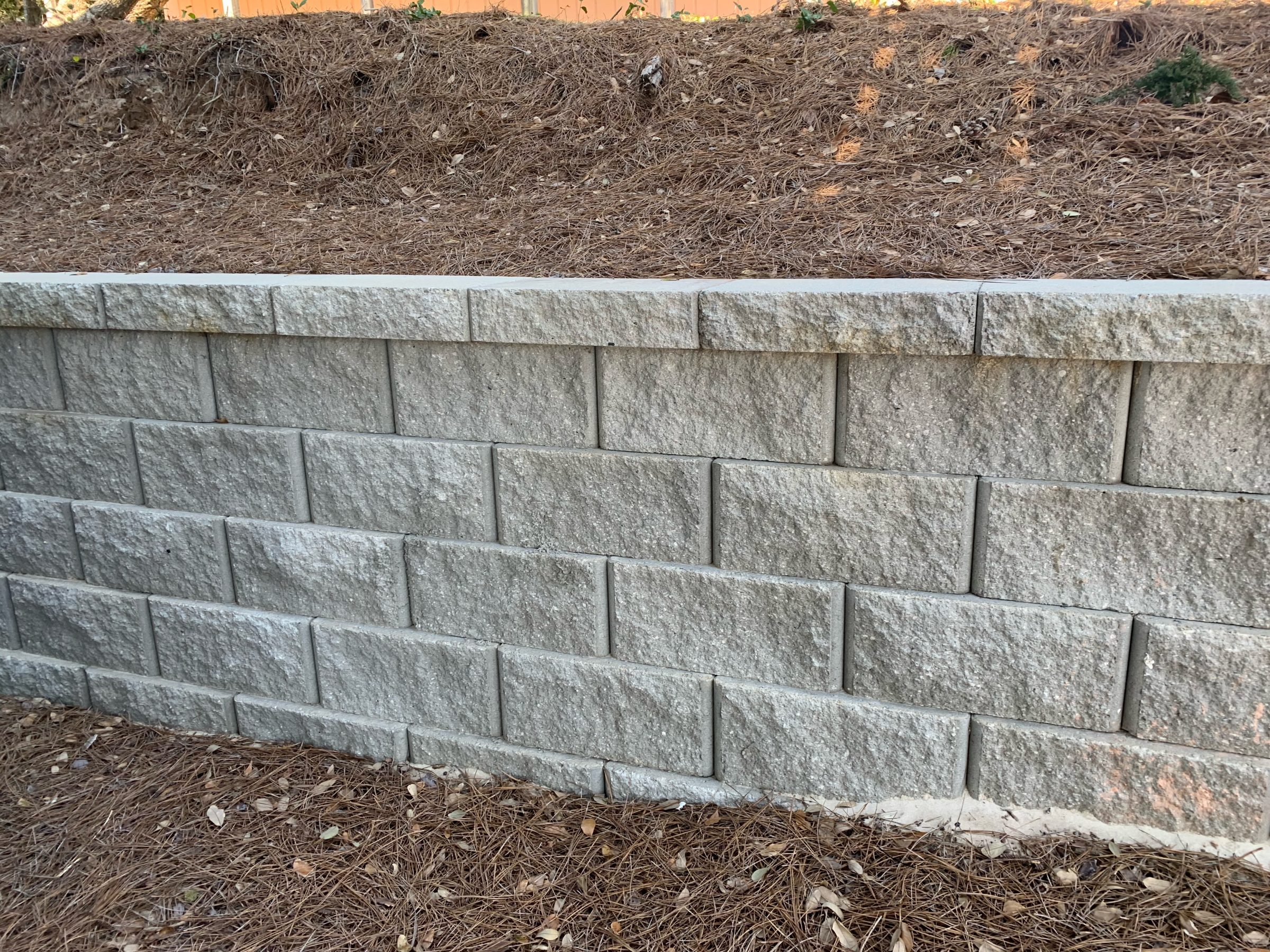 Gray stone retaining wall with pine needles on ground.
